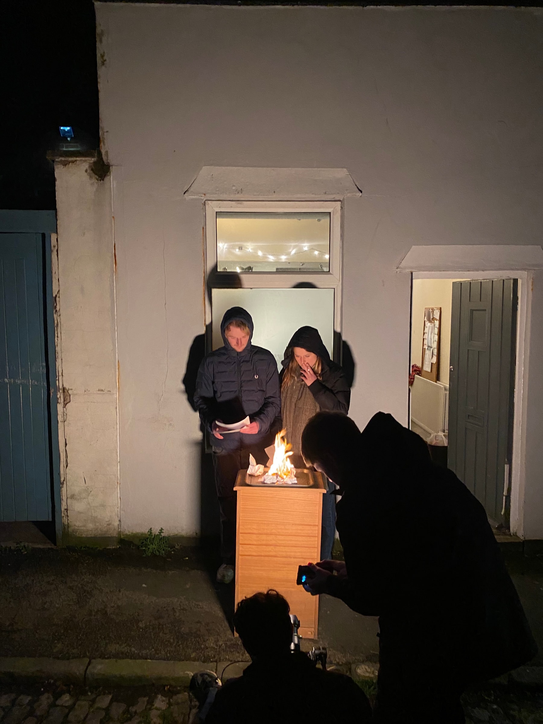 A night scene being filmed around a small fire outside a house